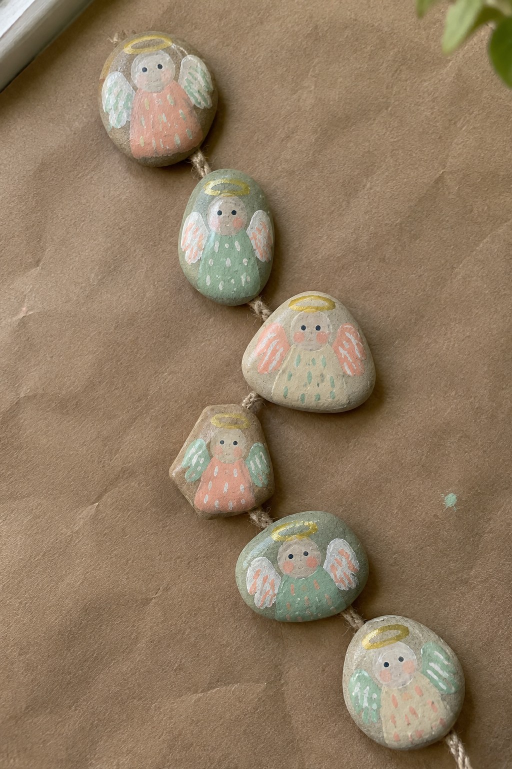 Six painted stone angels with halos and wings strung on twine over brown paper.