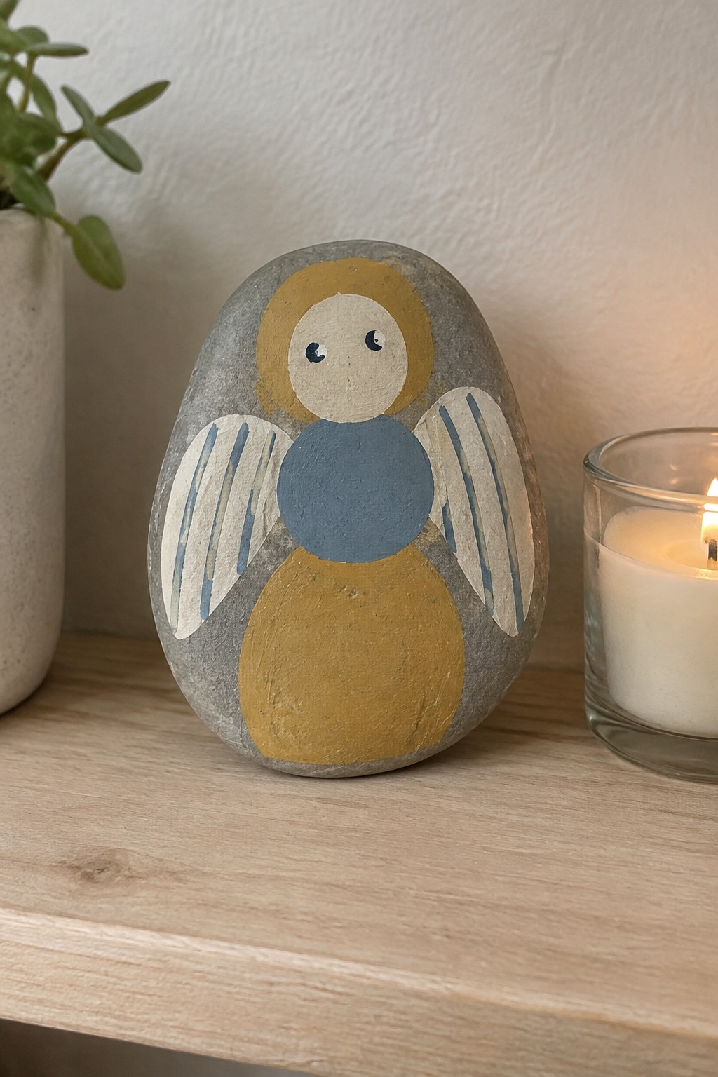 Painted gray rock angel with yellow halo, blue body, striped wings near candle