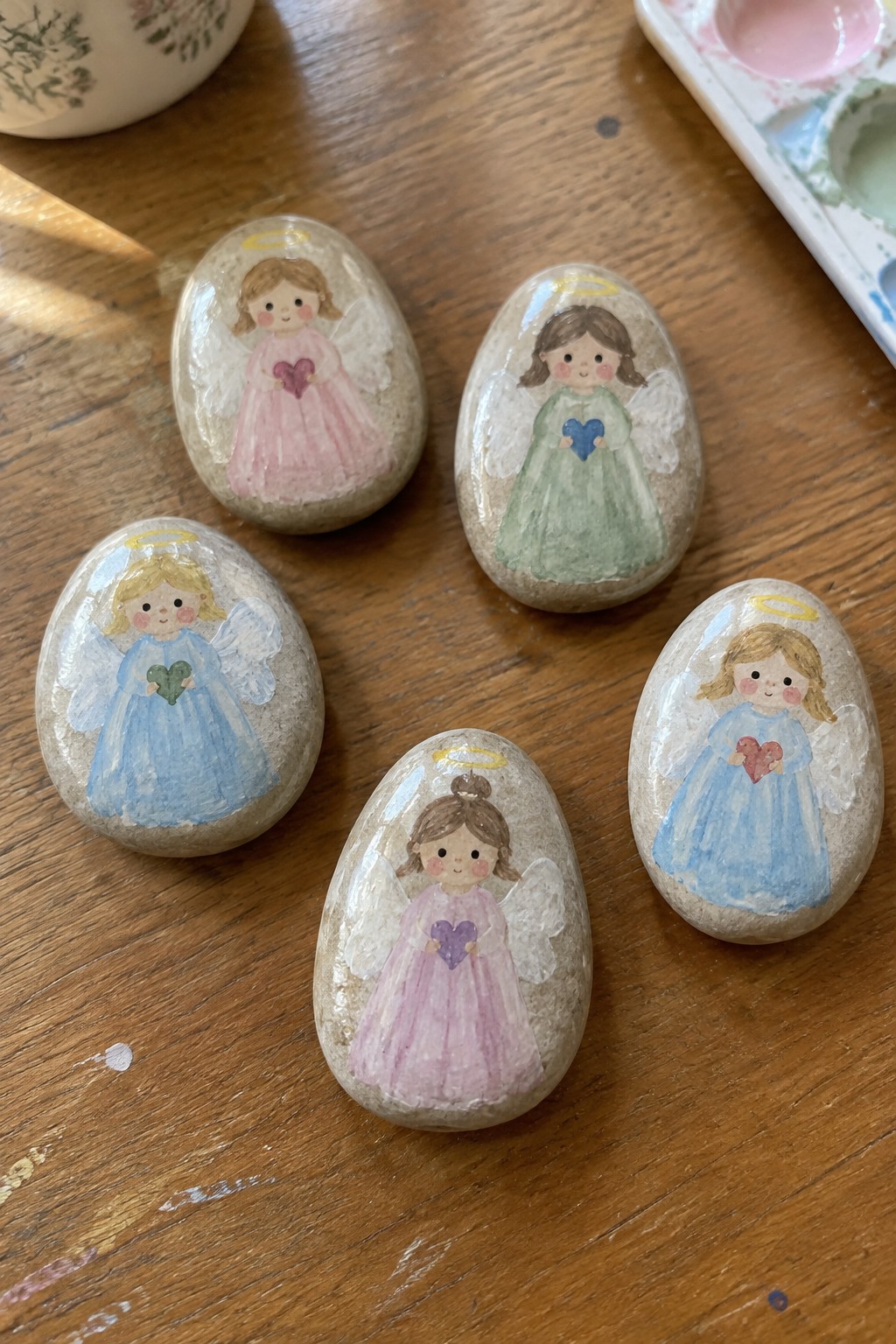 Five painted angel rocks on wood, girls in pastel dresses with halos and hearts.