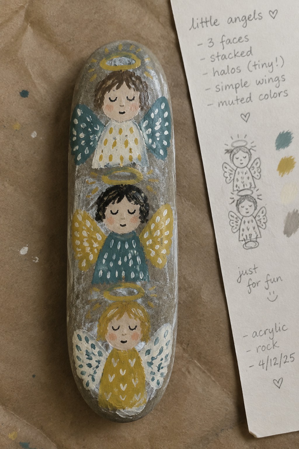 Three stacked angels with halos and dotted wings painted on an oval rock