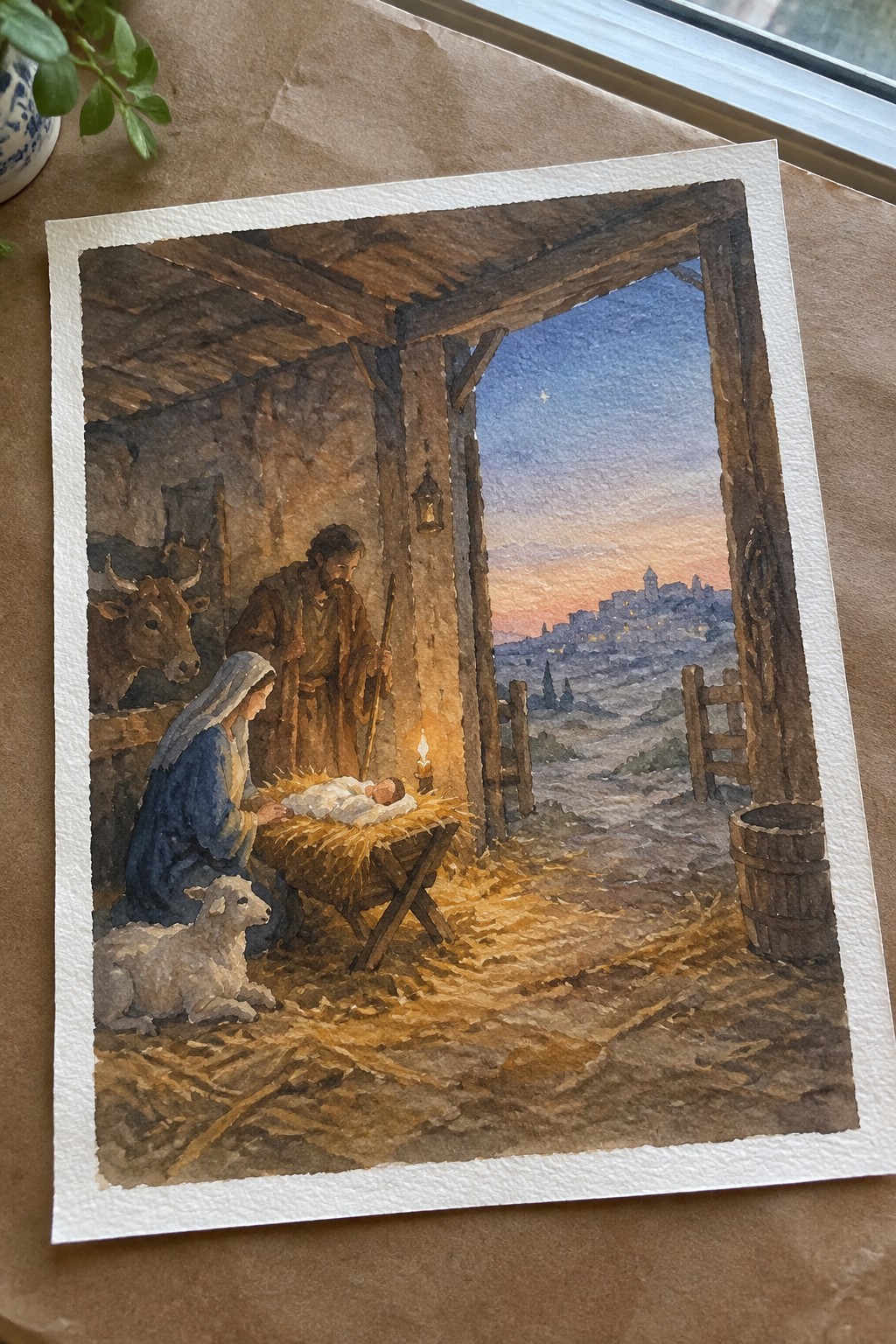 Watercolor nativity: Mary in blue kneels by manger with baby Jesus, Joseph with staff, lamb and ox.