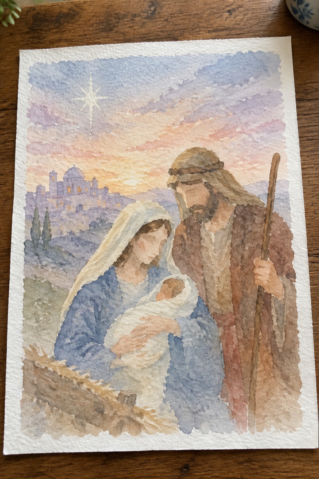 Watercolor of Mary in blue holding swaddled infant, robed Joseph with staff, star over Bethlehem.
