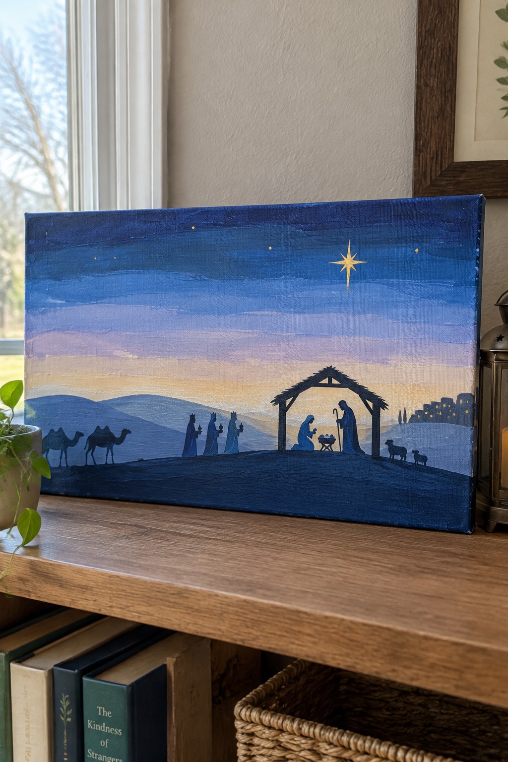 Canvas painting of a nativity silhouette scene with figures, camels, stable, and large star against a blue-to-peach gradient sky on a wooden shelf.