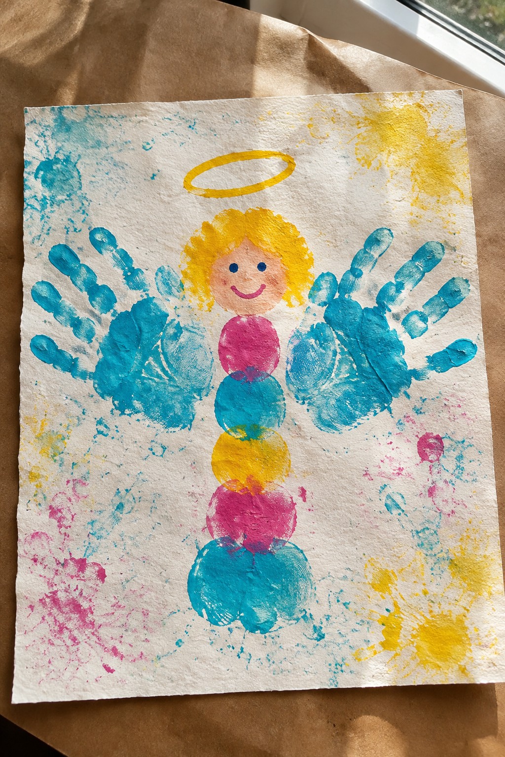 Handprint angel with blue wings, yellow halo, and multicolored stacked circle body amid paint splatters