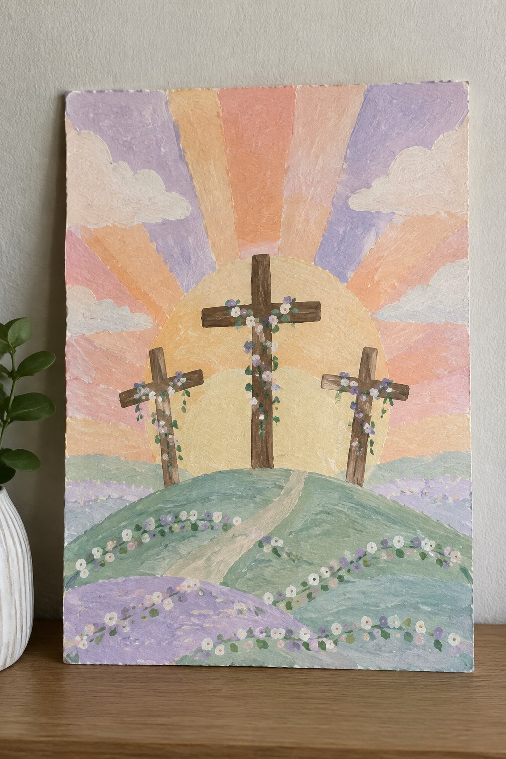 A painting of three wooden crosses on green and purple hills with flowers, set against a large yellow sun and pastel rays in orange, pink, and purple.