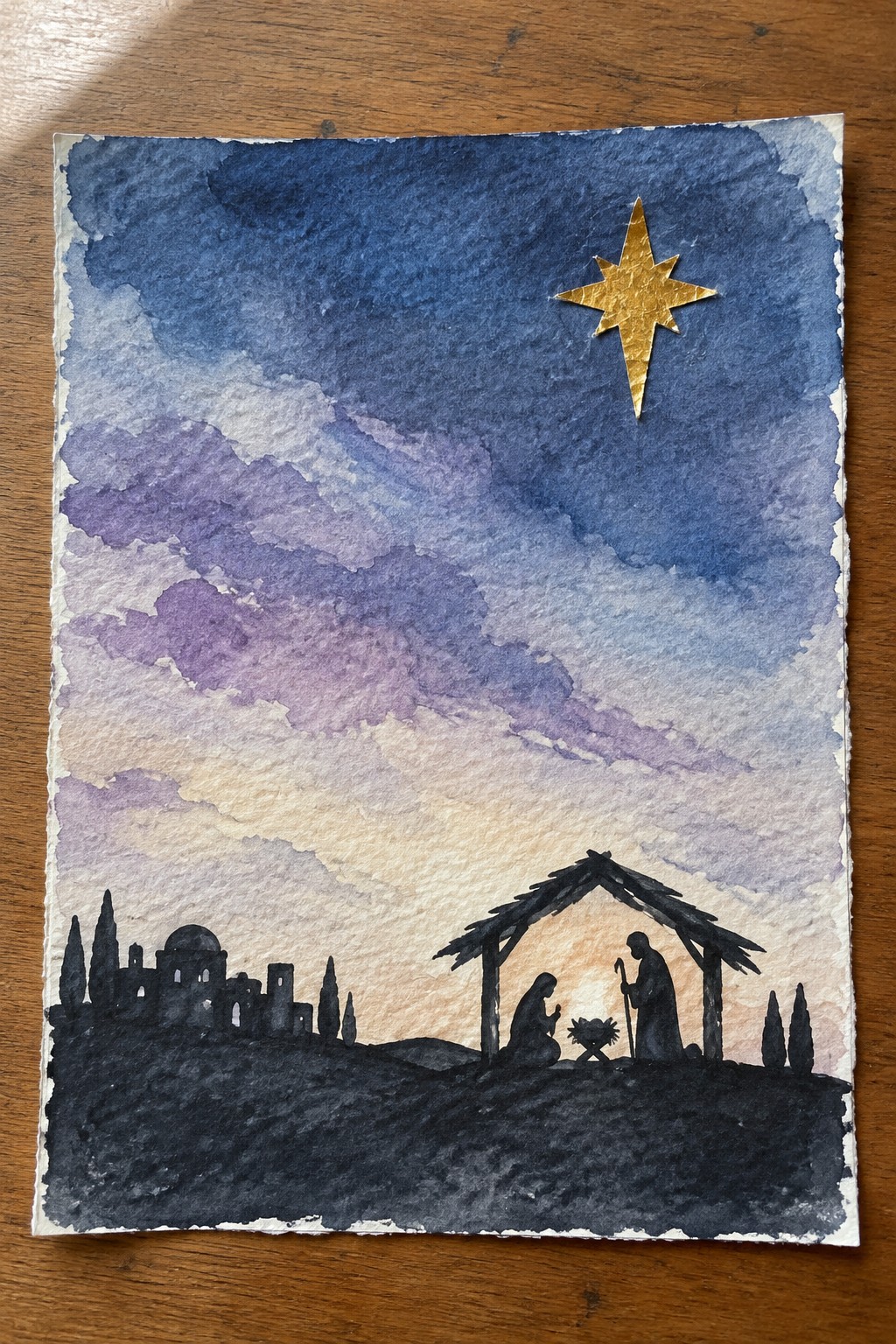 Watercolor nativity scene with golden star above silhouetted stable, kneeling Mary, and distant town.