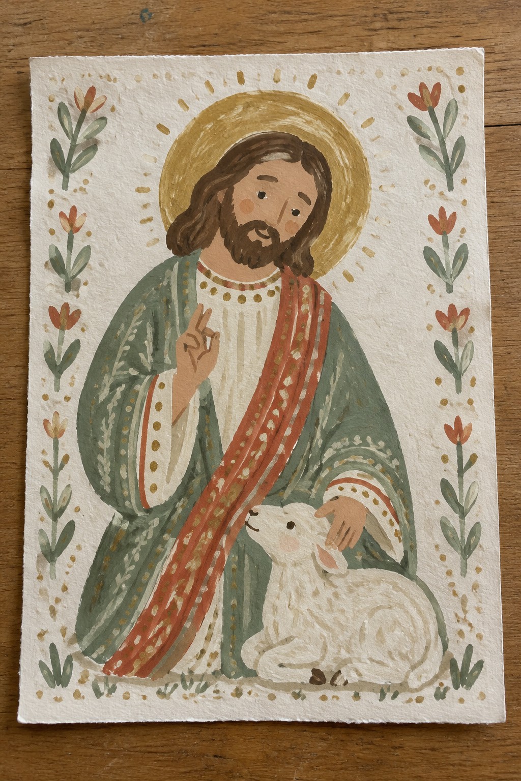 Watercolor painting of a haloed Jesus figure in green and red robes gently touching a white lamb, with floral borders on each side.