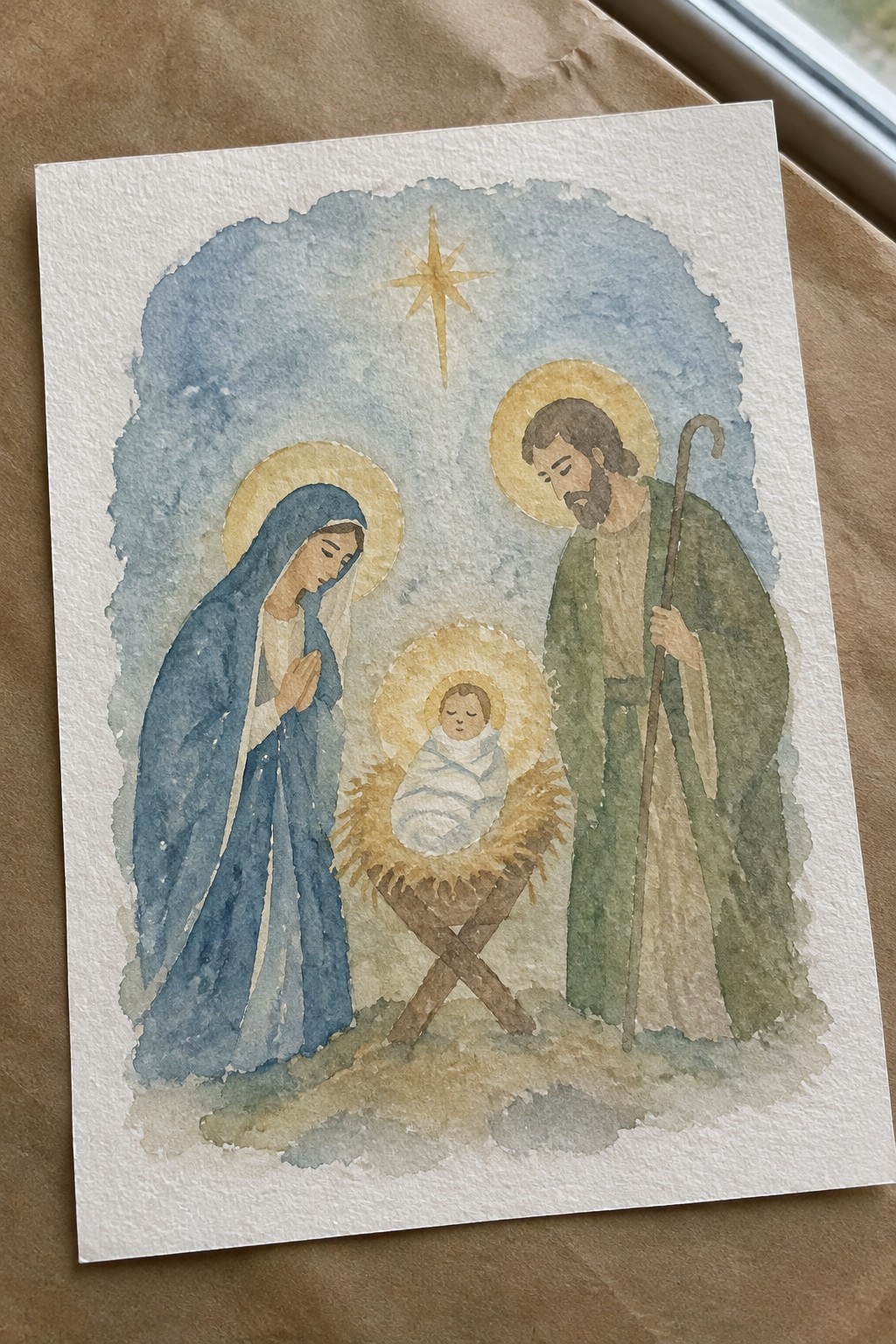 Watercolor nativity with Mary in blue, Joseph in green, baby Jesus in manger, star above.