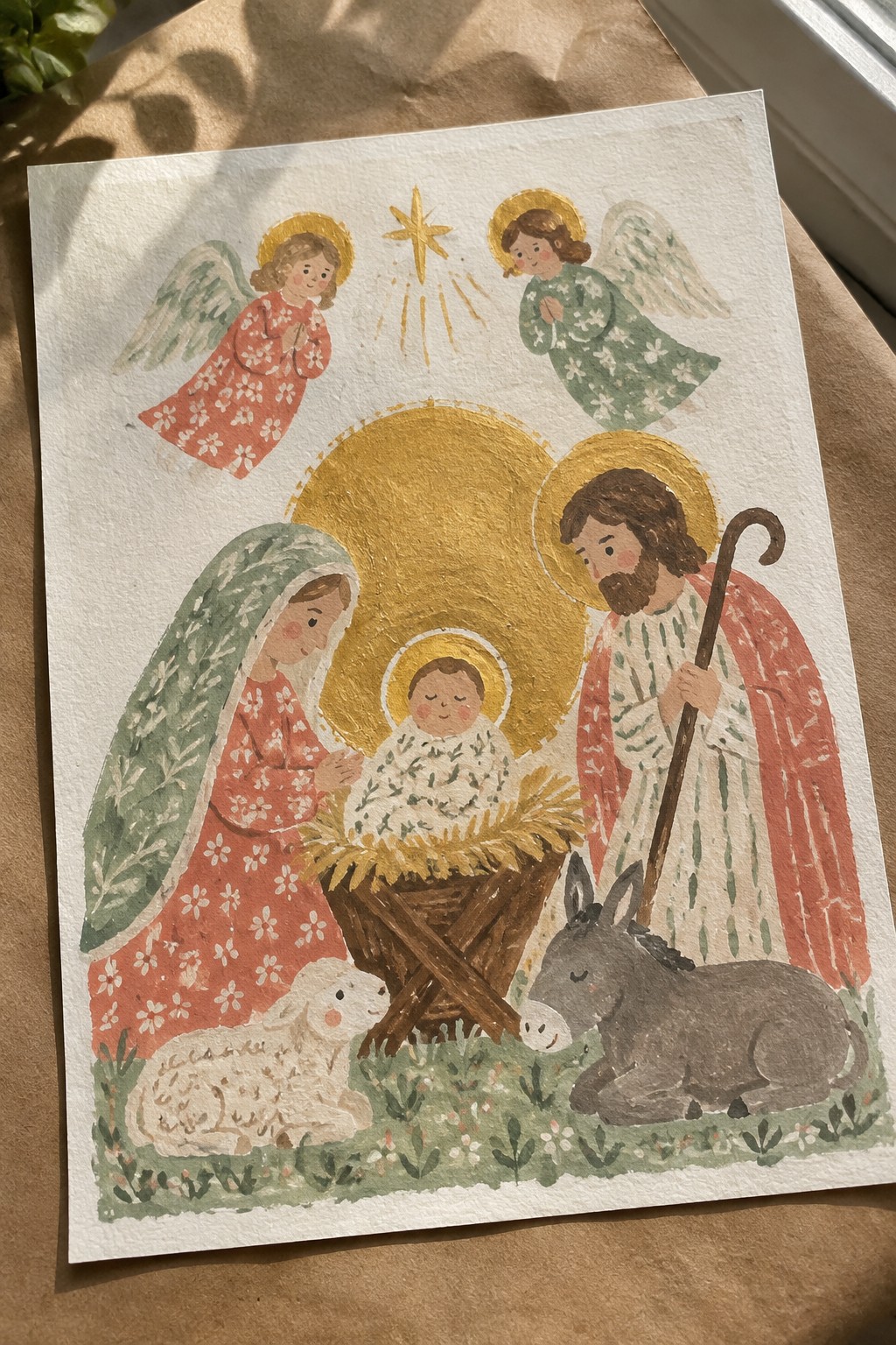 Watercolor nativity with Mary in red floral dress, Joseph, haloed baby, angels, star, sheep and donkey.