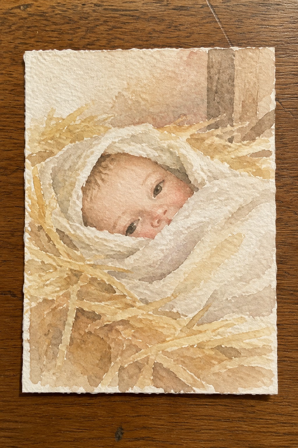 Watercolor of swaddled baby nestled in golden straw against wooden background