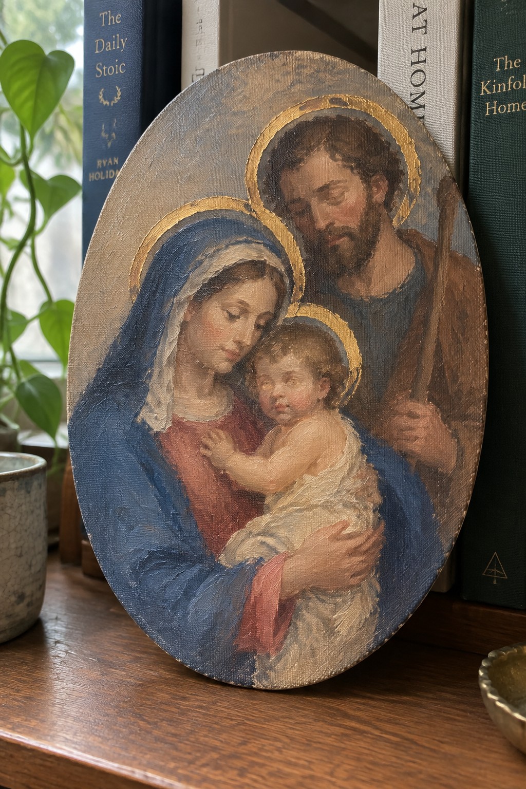Oval oil painting of haloed Mary in blue veil cradling infant Jesus beside staff-holding Joseph.