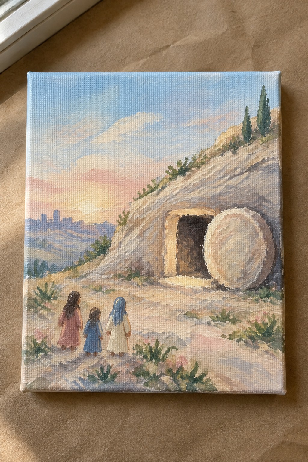 Three robed women approach an empty hillside tomb with a rolled-away stone at sunset.