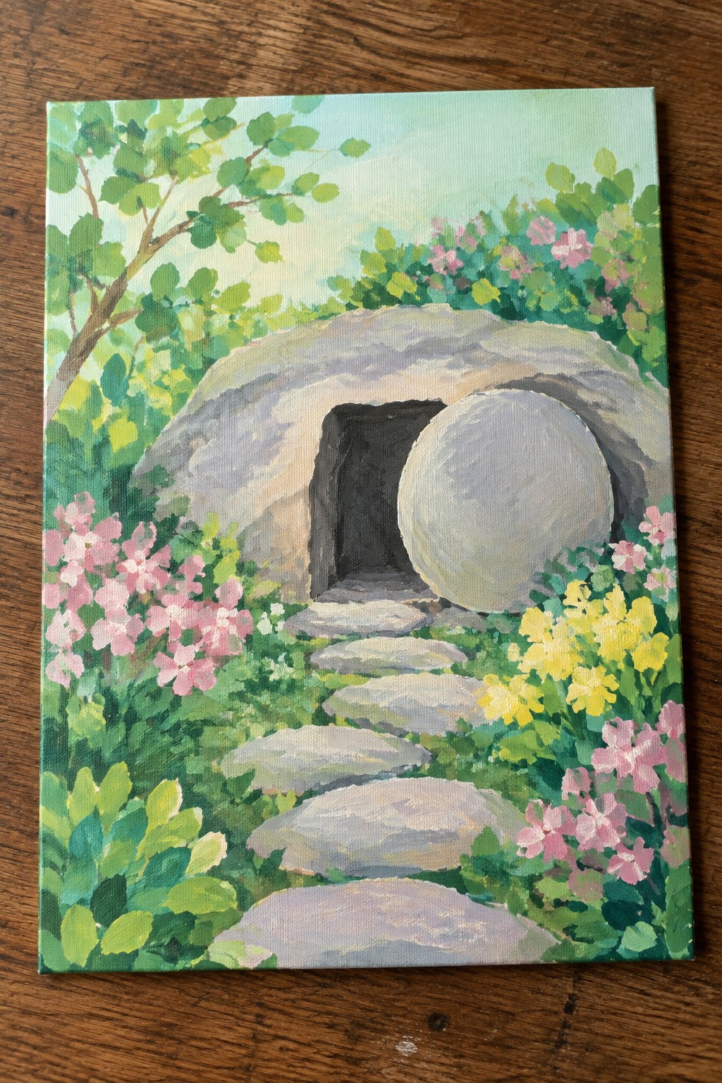 Open stone tomb with large boulder rolled aside, stone path amid pink and yellow flowers.