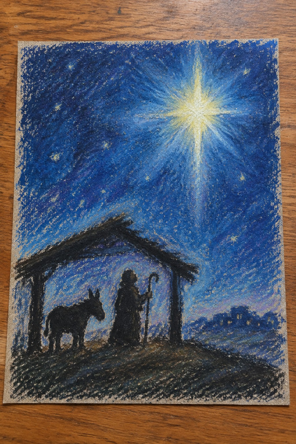 Chalk drawing of nativity with bright star, silhouetted stable, donkey, and robed figure.