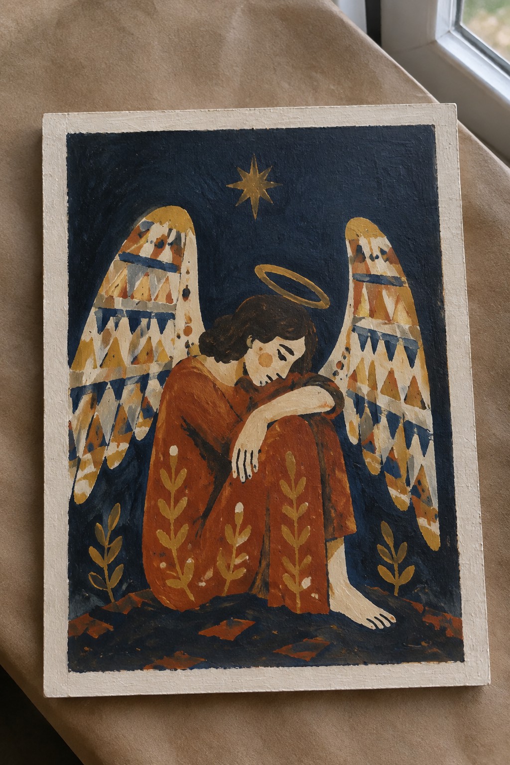Angel with halo and patterned wings sits sadly in brown robe under single star