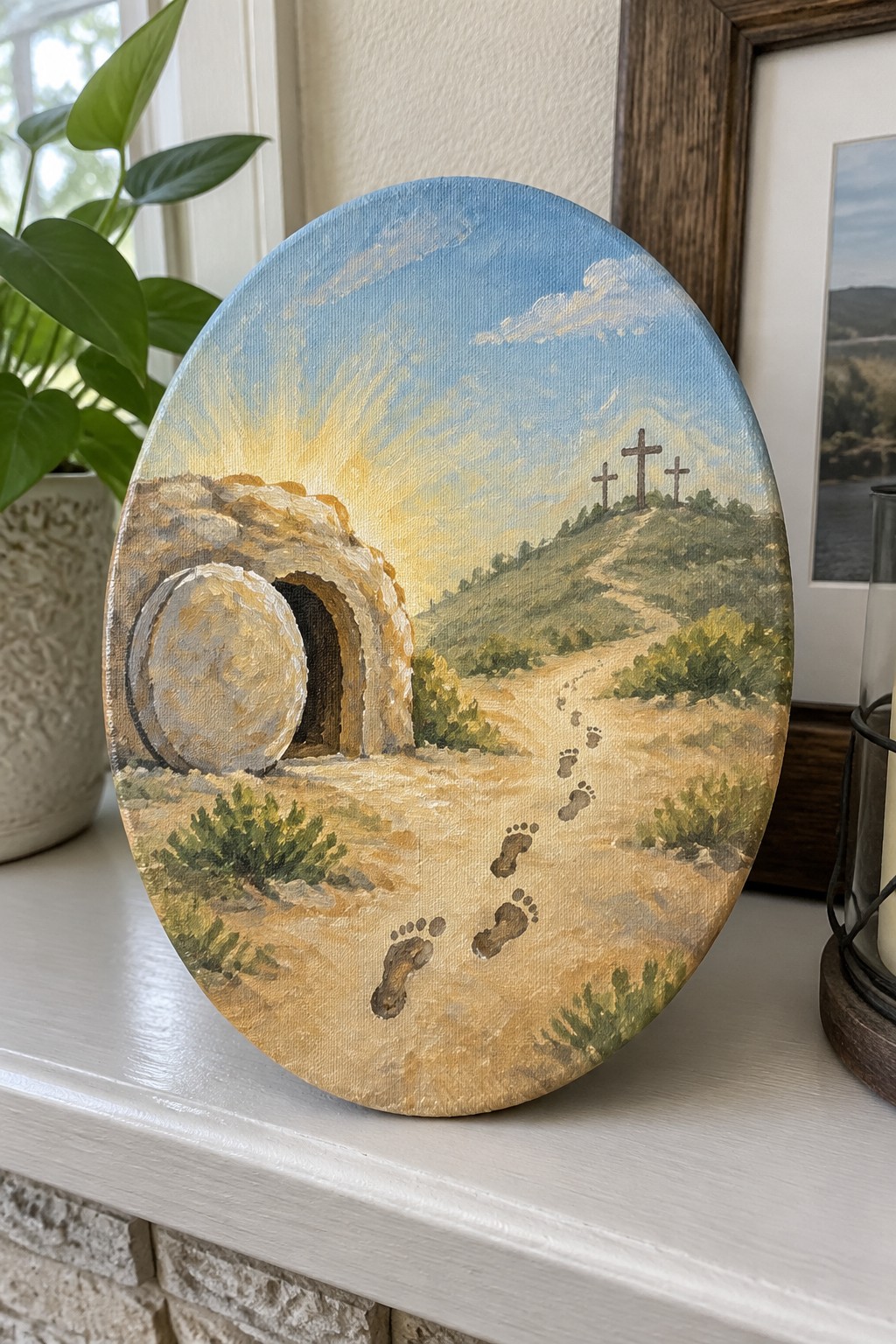 Oval canvas painting of empty tomb, footprints, and three crosses on hill at sunrise