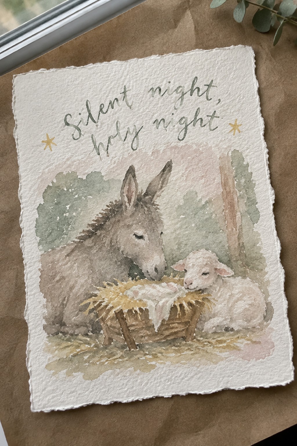 Watercolor nativity with donkey, lamb, and swaddled baby in manger under starry text.