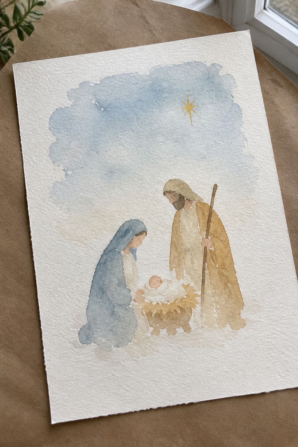 Watercolor nativity: Mary in blue kneels beside Joseph in brown with staff and baby in manger under star.