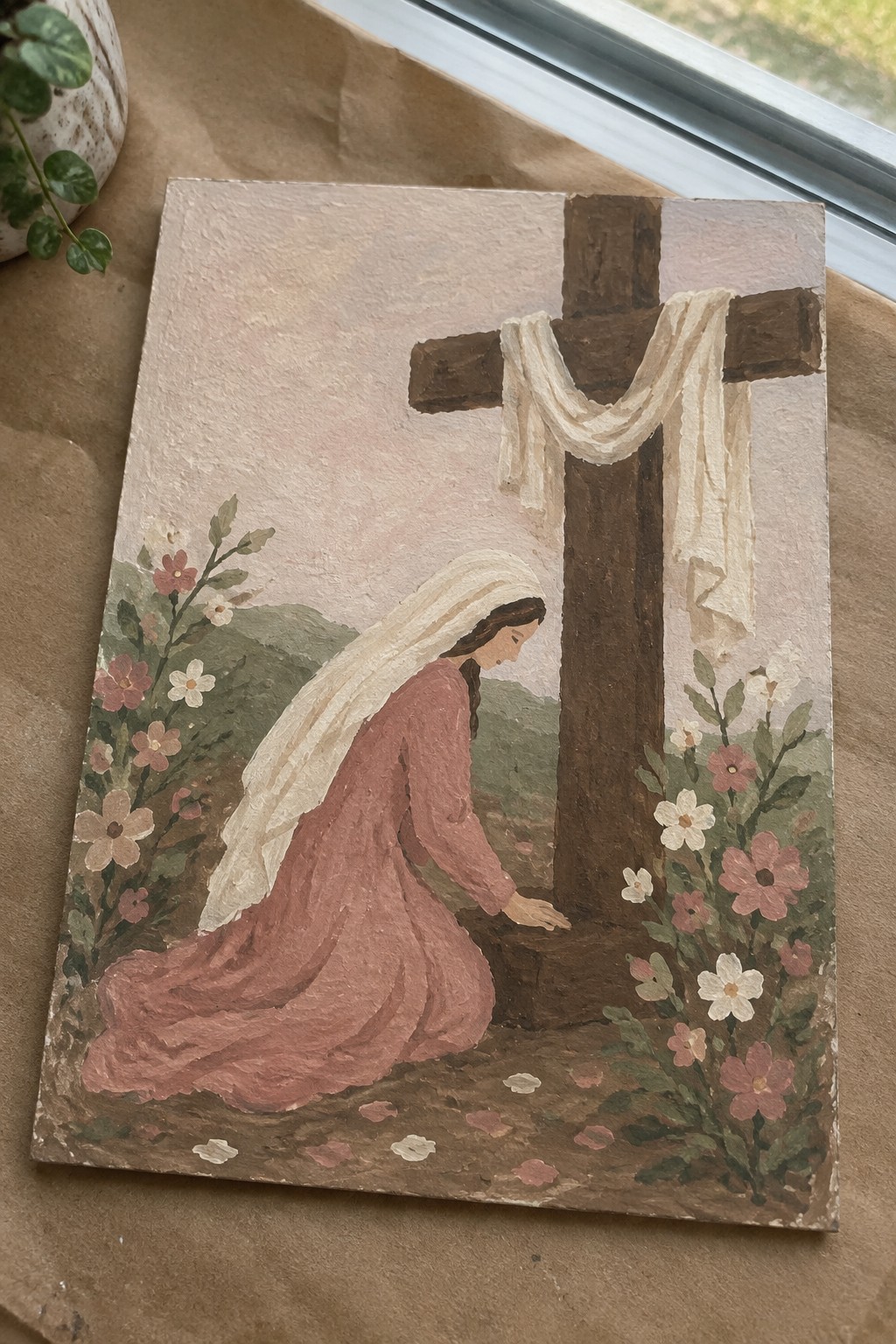 Woman in pink dress and white veil kneels before wooden cross with draped cloth amid flowers