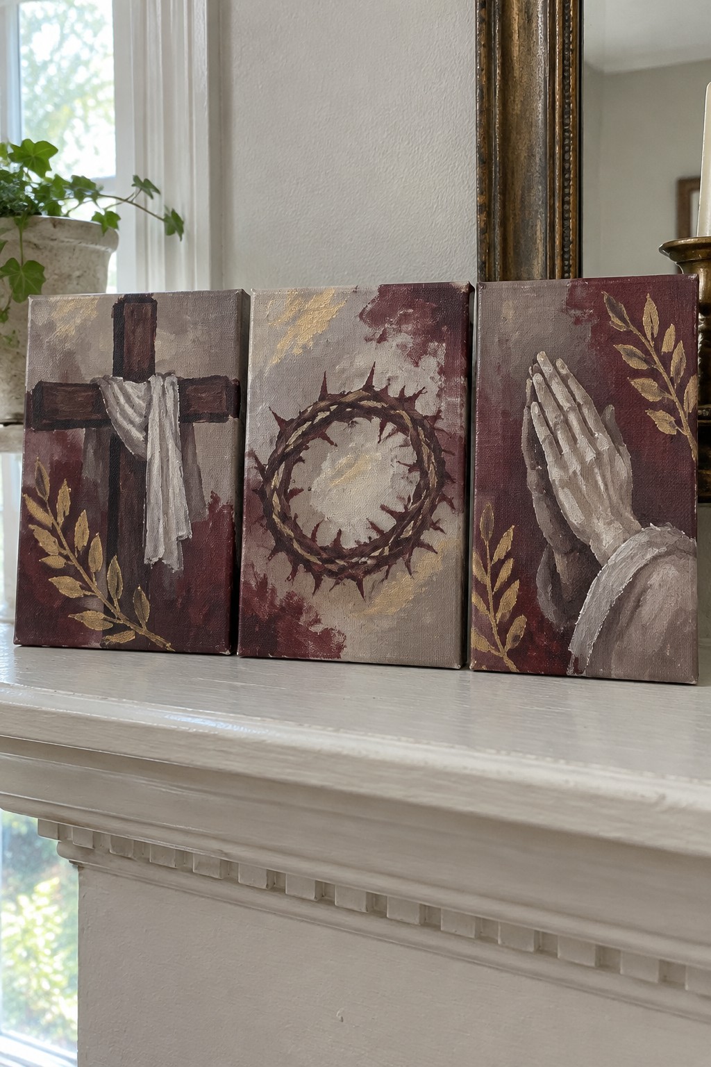 Three maroon-and-gold paintings on white mantel: cross, crown of thorns, praying hands.