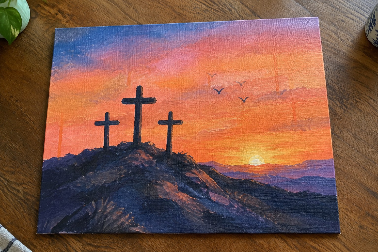 Three crosses on a hill under vibrant orange-pink sunset sky with flying birds.