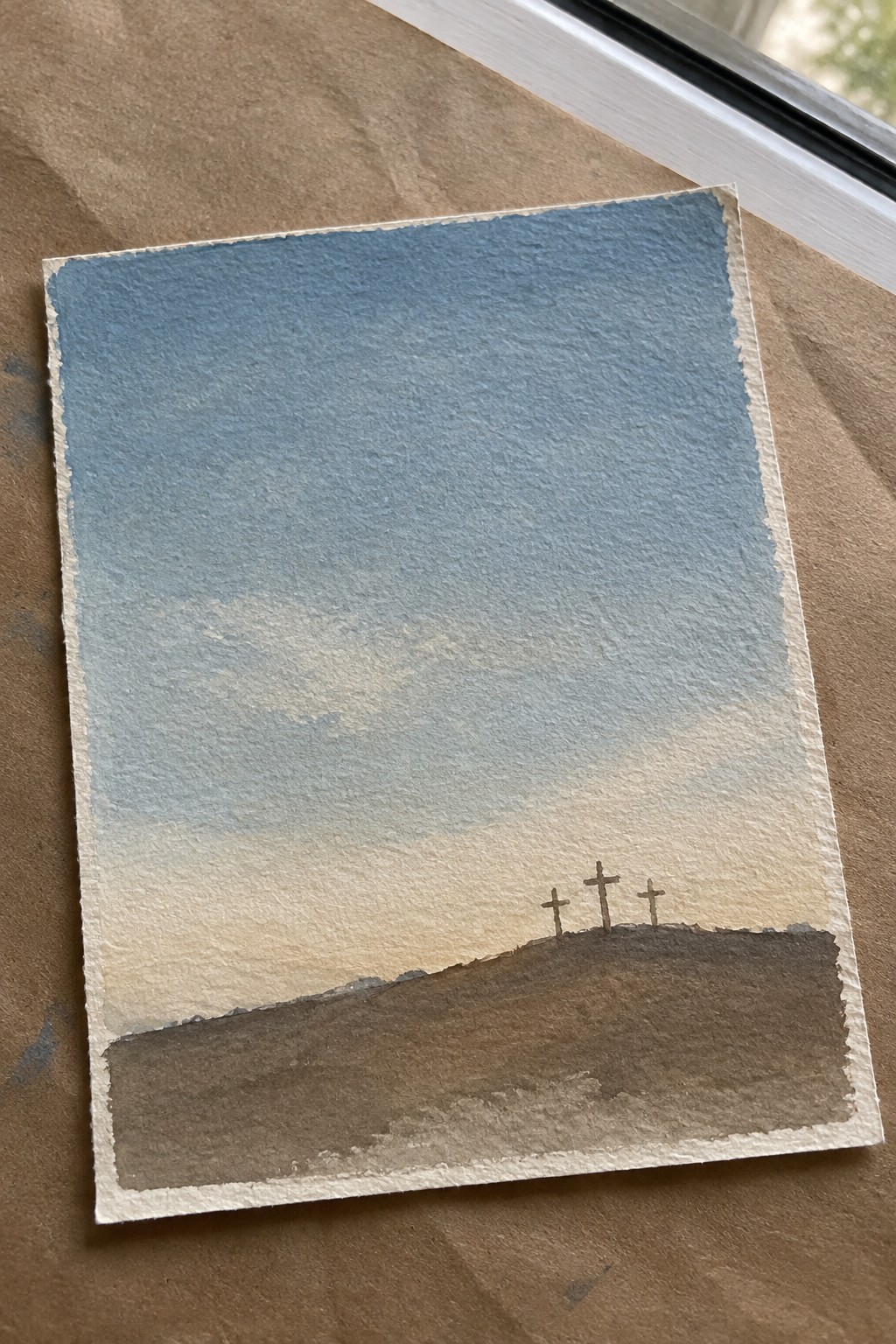 Watercolor painting of three crosses on a brown hill under blue sky