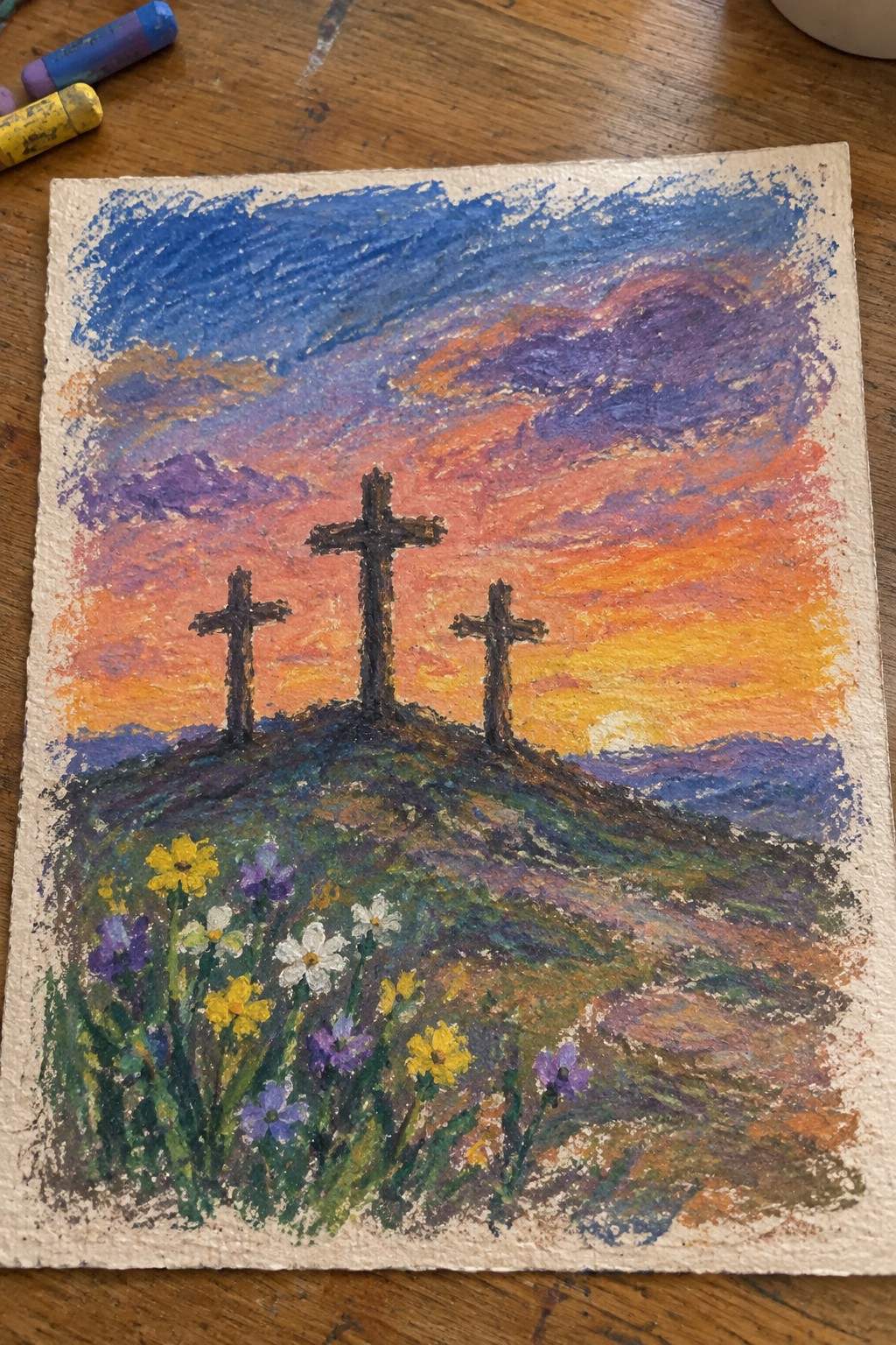 Chalk pastel of three crosses on a hill under vibrant orange-purple sunset with wildflowers.