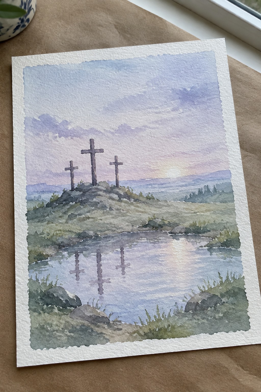 Three crosses on a hill reflected in a pond under a pastel sunset sky in watercolor