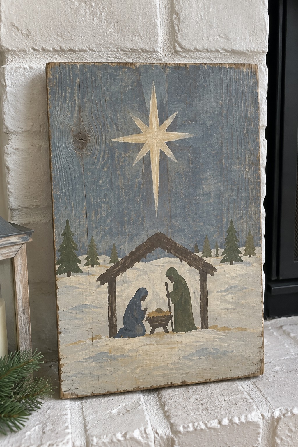 A painted nativity scene on a distressed wooden panel showing a star above a stable with Mary, Joseph, and baby Jesus in a snowy landscape with pine trees.