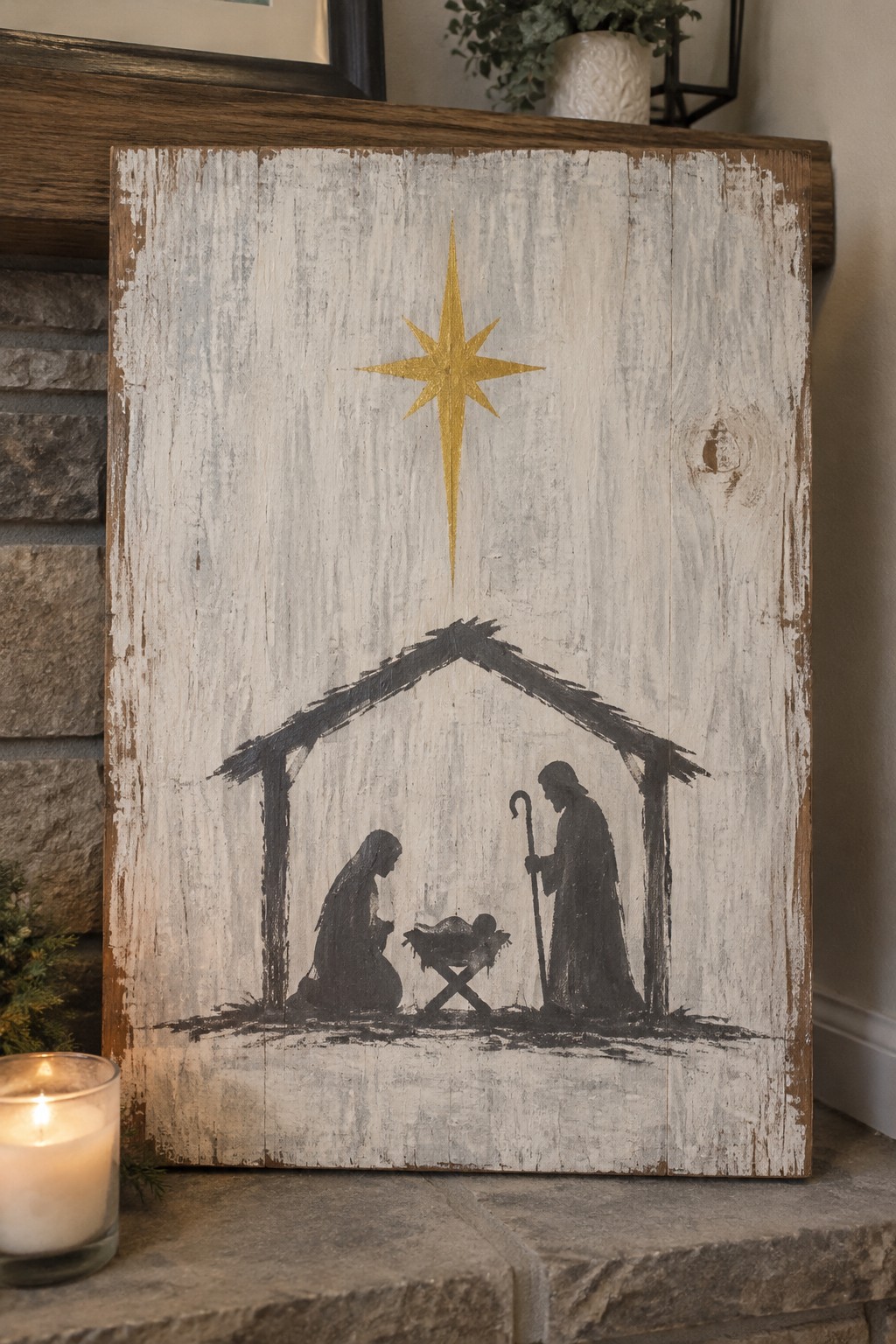 A whitewashed wooden panel shows a black silhouette nativity scene with a golden star above the stable, sitting on a stone surface beside a lit candle.