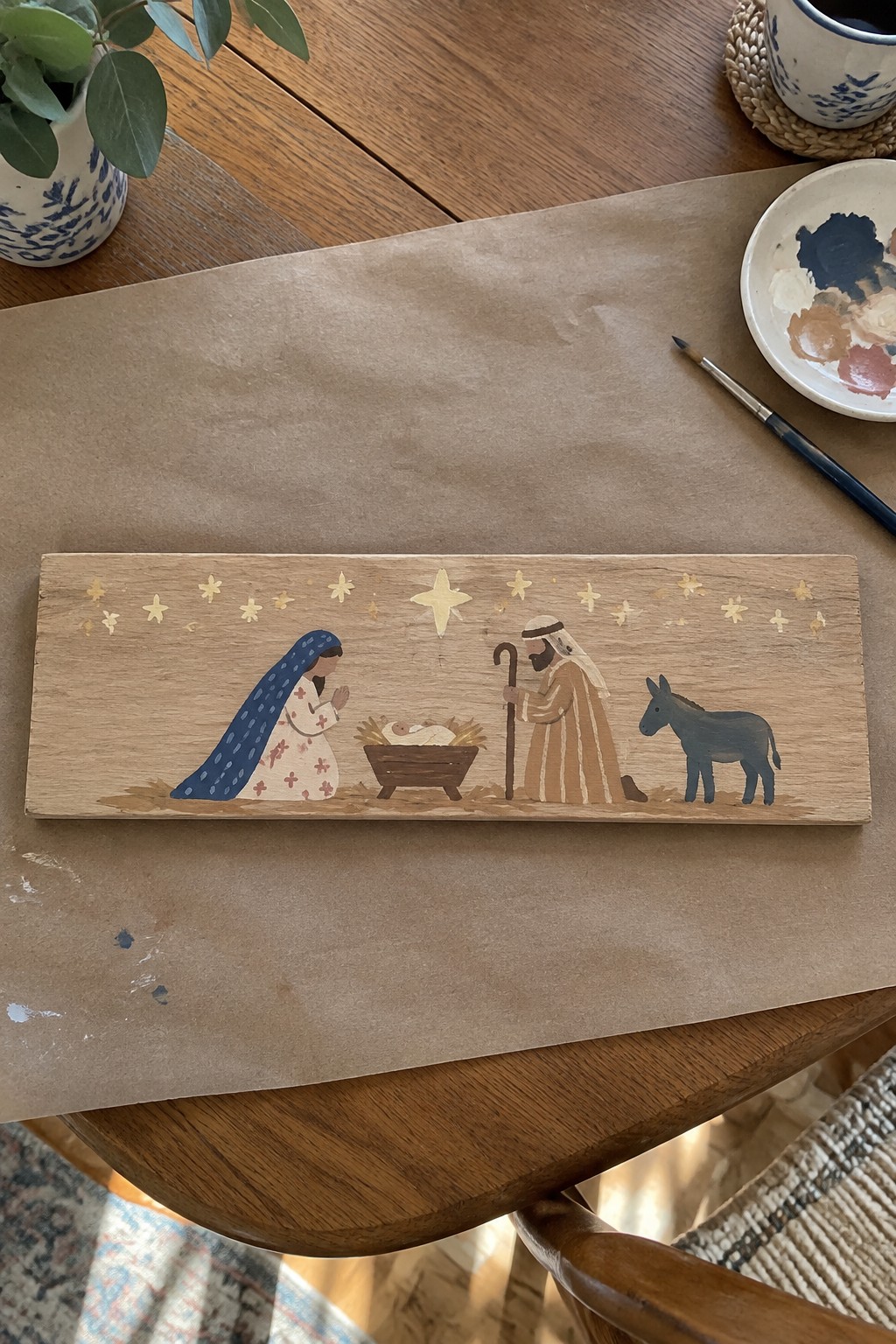 Wooden plaque with painted nativity: Mary in blue veil, Joseph in stripes, donkey, golden stars.