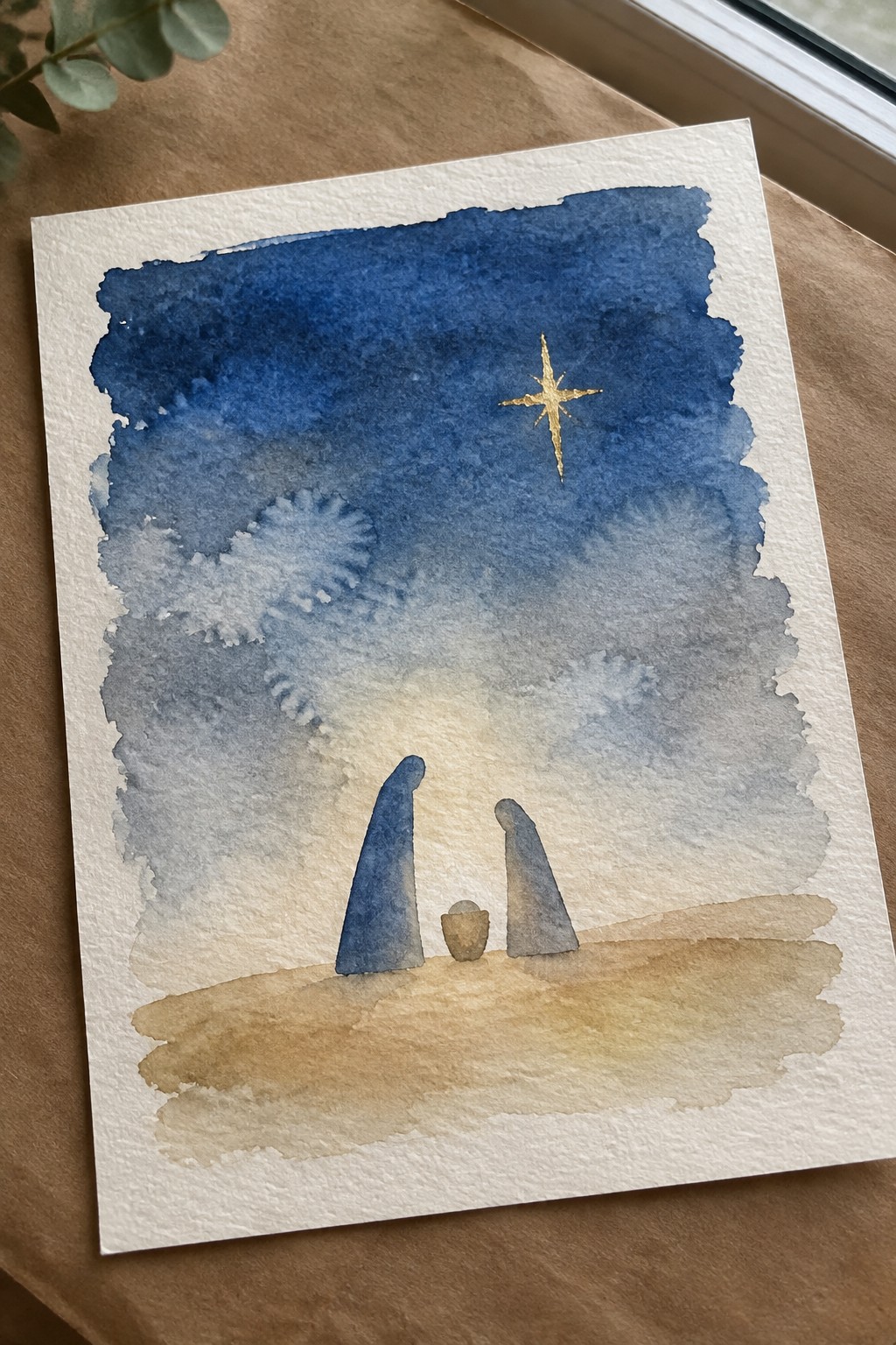 Watercolor nativity with two blue-robed figures, manger, and golden star in blue sky.