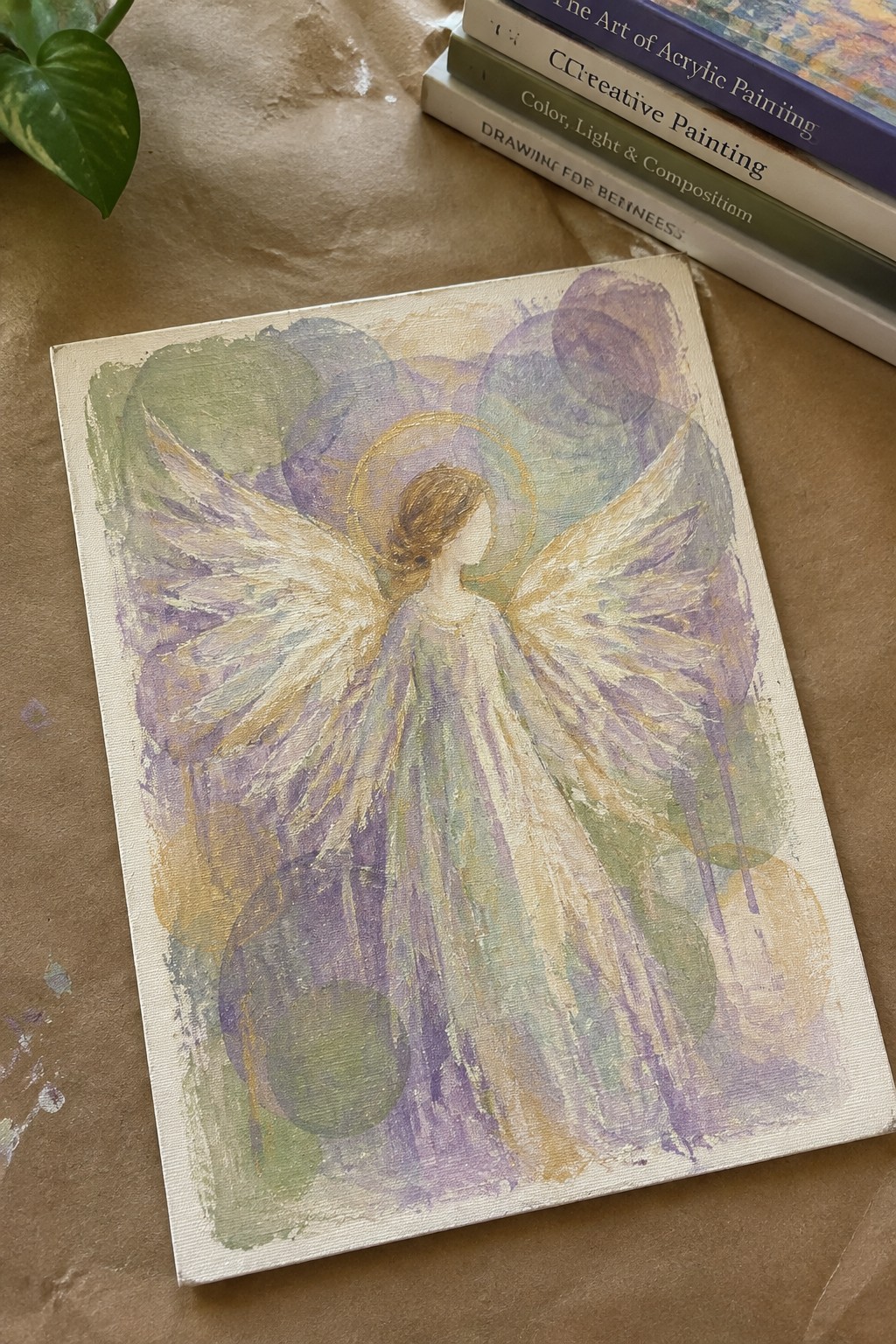 Soft pastel angel painting with golden halo and flowing wings on canvas beside books