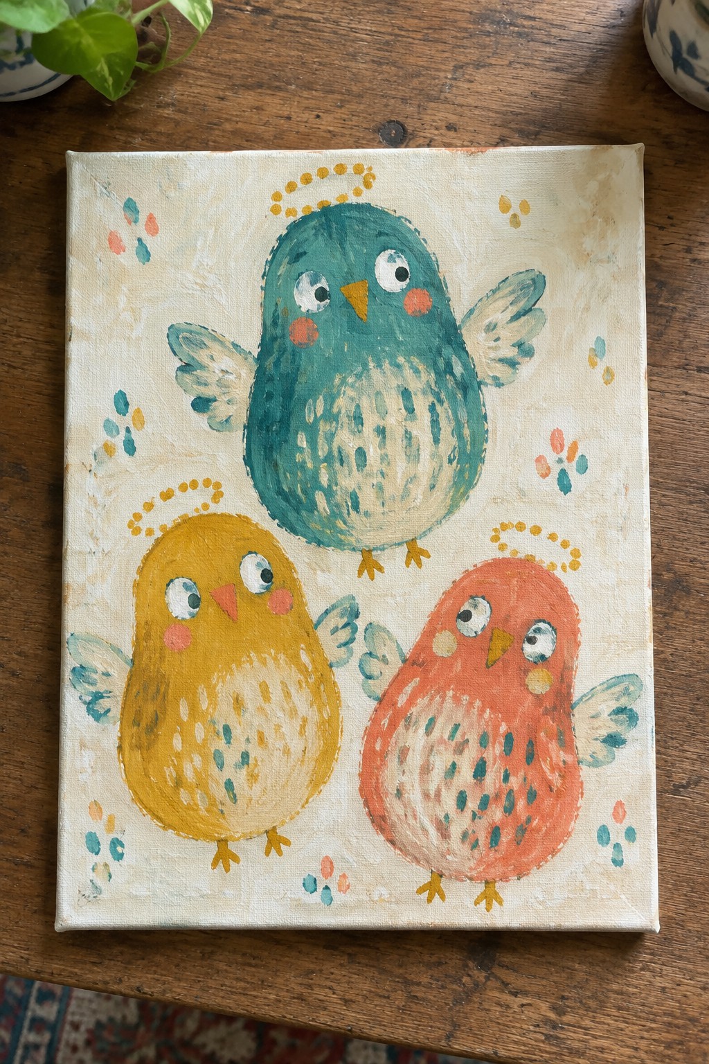 Three round birds with dotted halos painted on a canvas, one teal above two smaller yellow and orange birds.