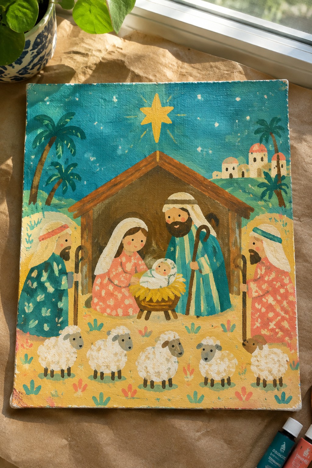 Vibrant folk-art nativity painting with robed figures, manger, sheep, star, and palm trees.