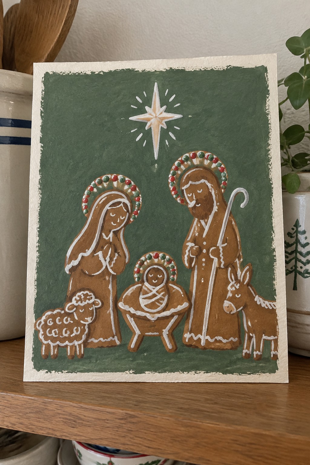 Gingerbread-style nativity with haloed Mary, Joseph, baby Jesus, sheep, donkey, and star on green.